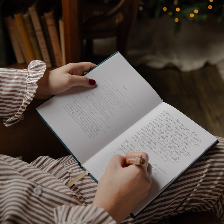 A person in a brown and white striped pajama shirt holds an open notebook or journal, writing in a handwritten page with a pen. The scene is set in a cozy, dimly lit room with a blurred background of books and warm lights.
