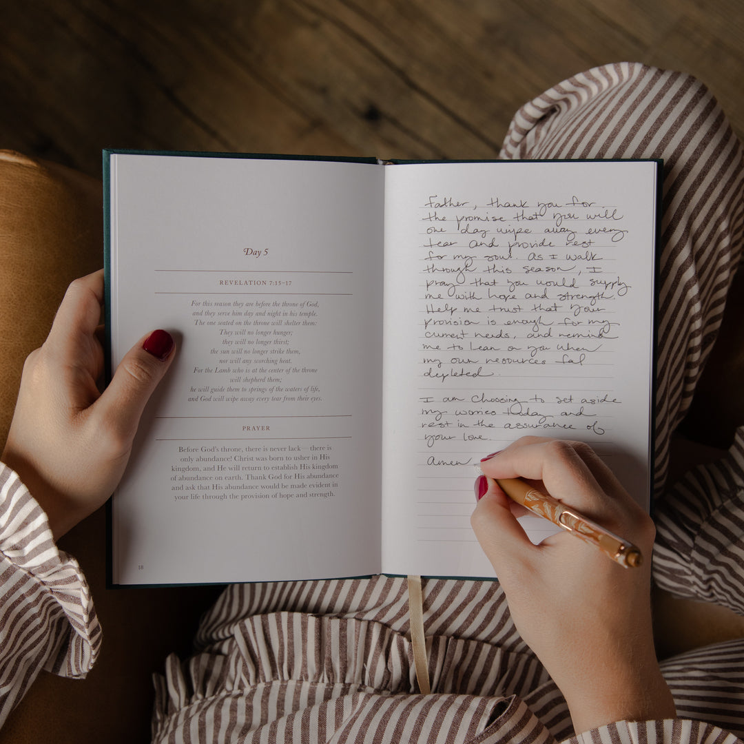 An open journal or diary lying on a striped bed, with a person's hands holding the pages and writing in the right-hand column. The person is wearing a brown and white striped shirt, and the background shows a cozy bedroom setting with a wooden floor.