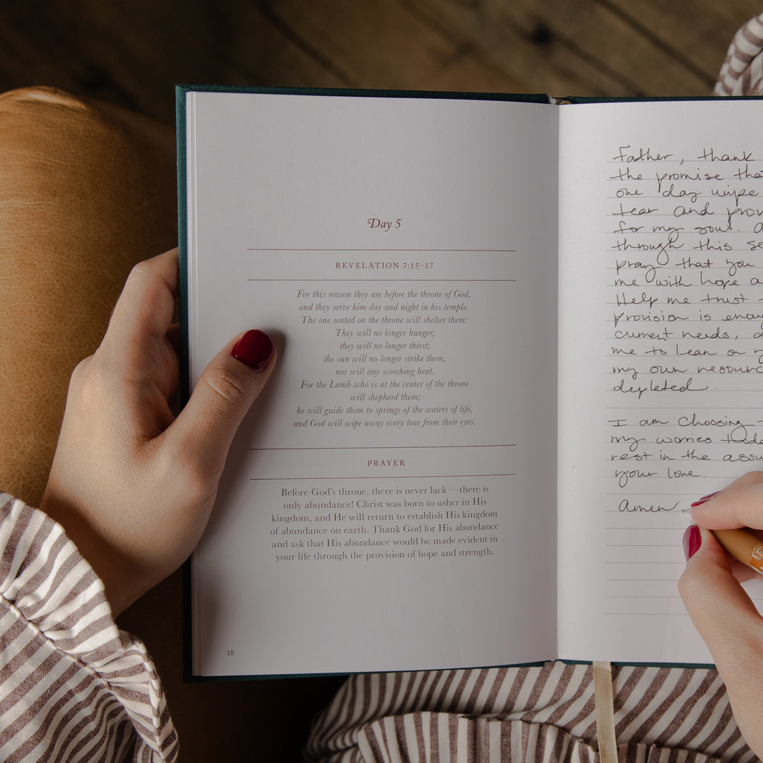 An open book showing handwritten and printed text from Revelation 5:1-11, held open by two hands with red painted fingernails. The person is wearing a brown and white striped shirt, and the background shows a warm brown leather surface.