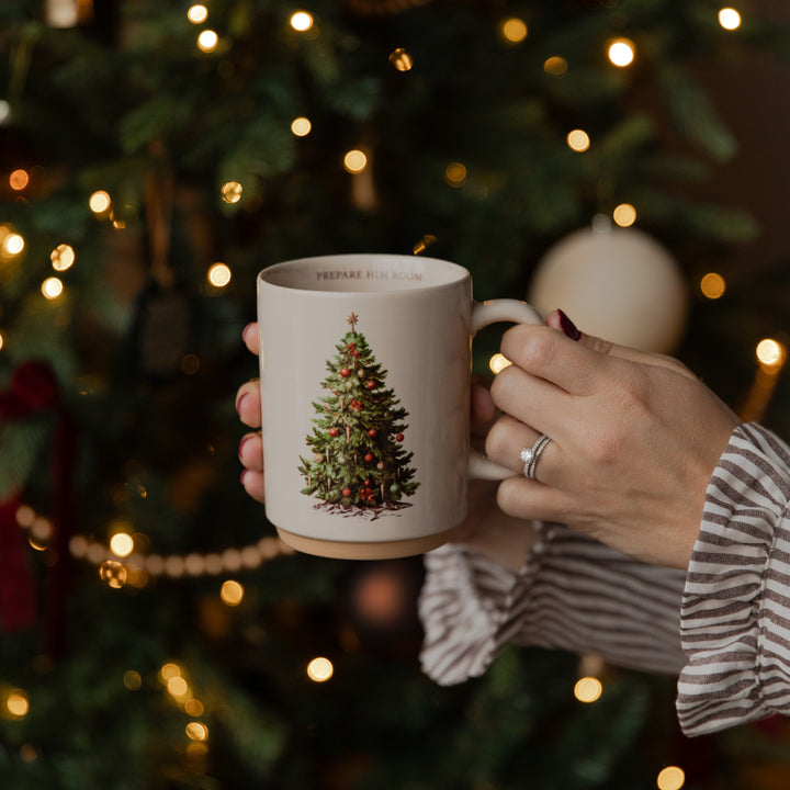 A person holds a ceramic mug featuring a vintage-style illustration of a Christmas tree with red ornaments, set against a festive, softly blurred Christmas tree background with warm lights.