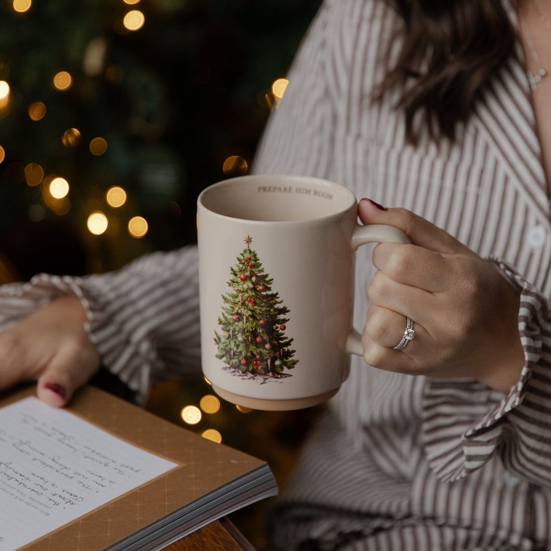 A person in a striped shirt holds a cream-colored ceramic mug featuring a vintage-style illustration of a Christmas tree. The background is softly blurred with warm bokeh lights, and an open notebook lies on the table nearby.