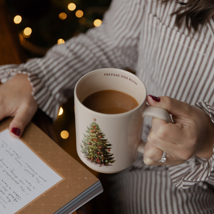 A person in a striped shirt holds a white ceramic mug with a Christmas tree illustration and the text 'PREPARE HIM ROOM' while sitting by a book with handwritten notes, with warm string lights blurred in the background.