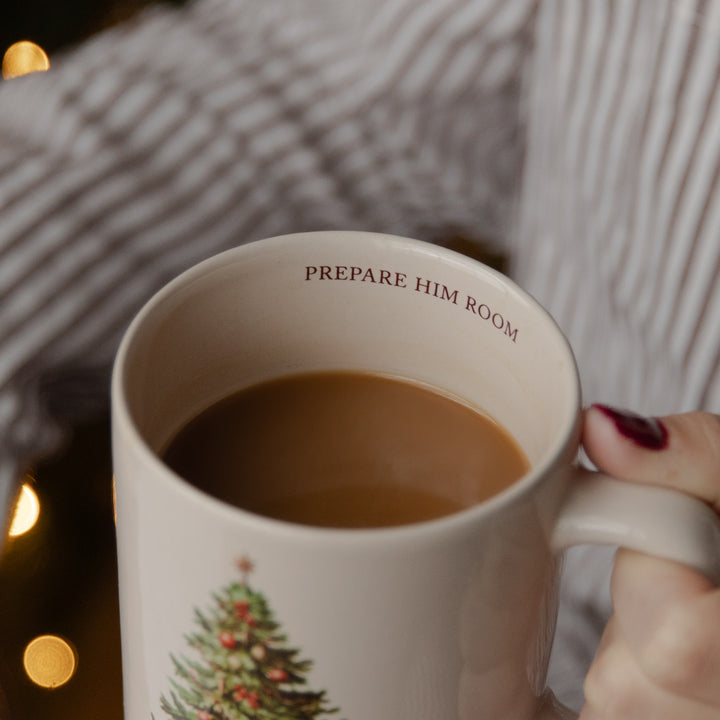 A close-up of a ceramic mug filled with a brown beverage, held by a hand with dark red nail polish. The mug is cream-colored with the text 'PREPARE HIM ROOM' printed inside the rim and a Christmas tree illustration near the bottom.