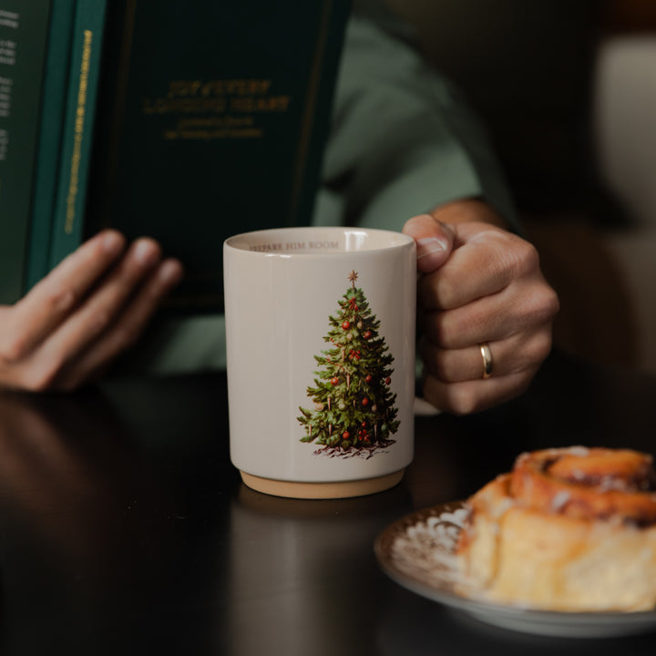 A ceramic mug with a Christmas tree illustration sits on a dark table, held by a person in a cozy indoor setting with a book and a pastry in the background.