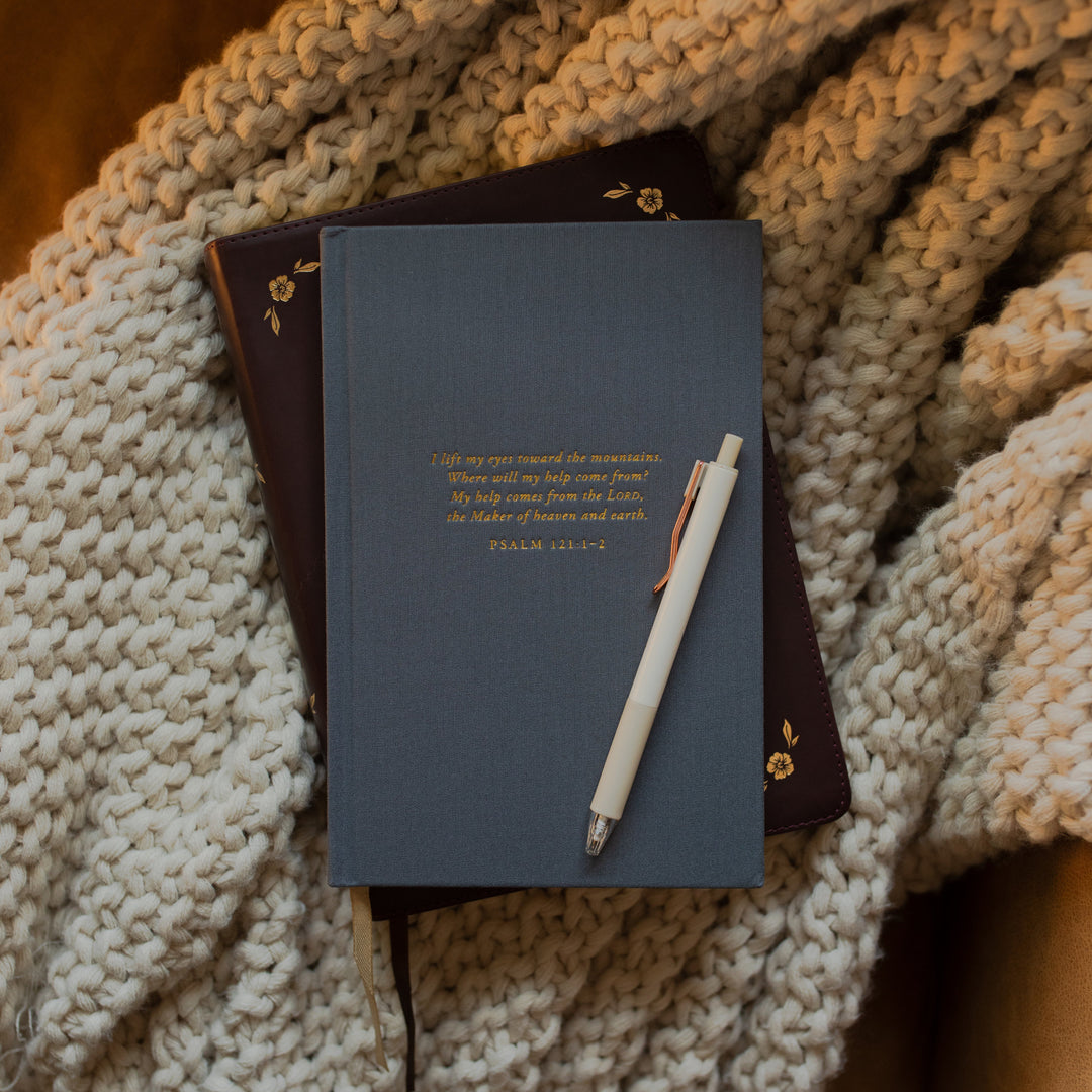 The Psalm 121 Dotted Journal cover on a closed bible with a pen resting on it while on a white knitted blanket