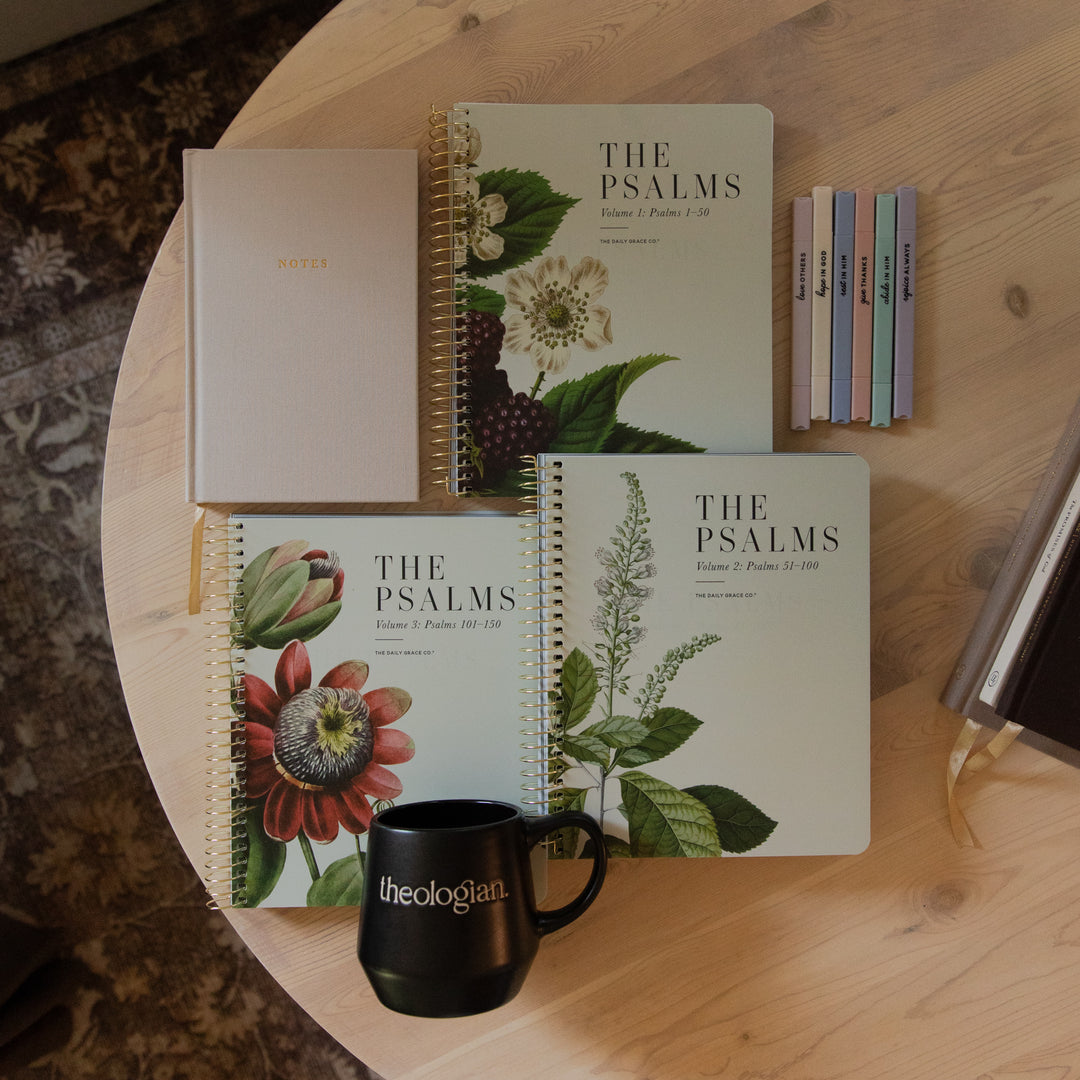 A styled flat-lay of The Psalms Bible set on a round wooden table, including a notebook, a black mug with 'theologian' printed on it, and a stack of colorful tabbed bookmarks.