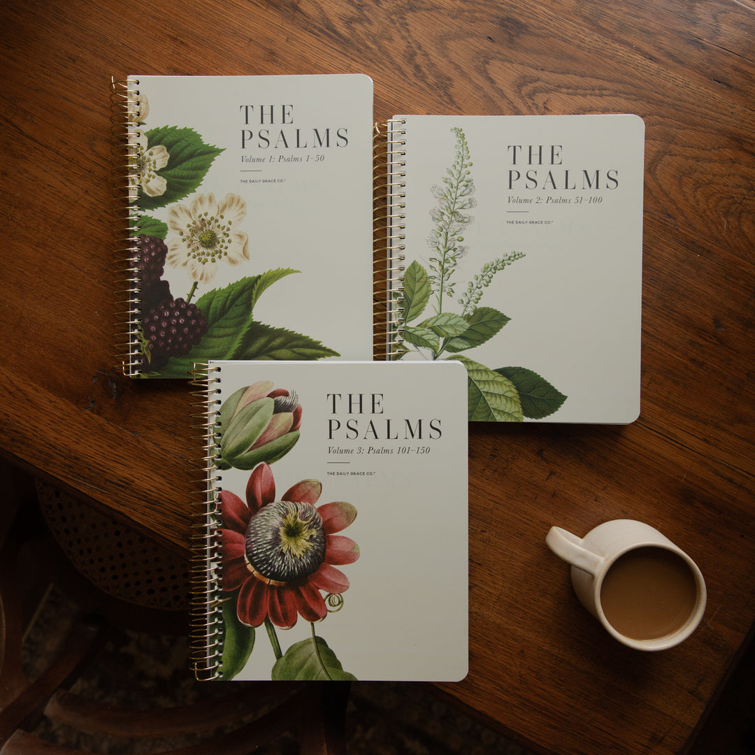 Three spiral-bound books titled "THE PSALMS" arranged on a wooden table, each with different botanical illustrations on the cover, plus a white mug of coffee nearby.