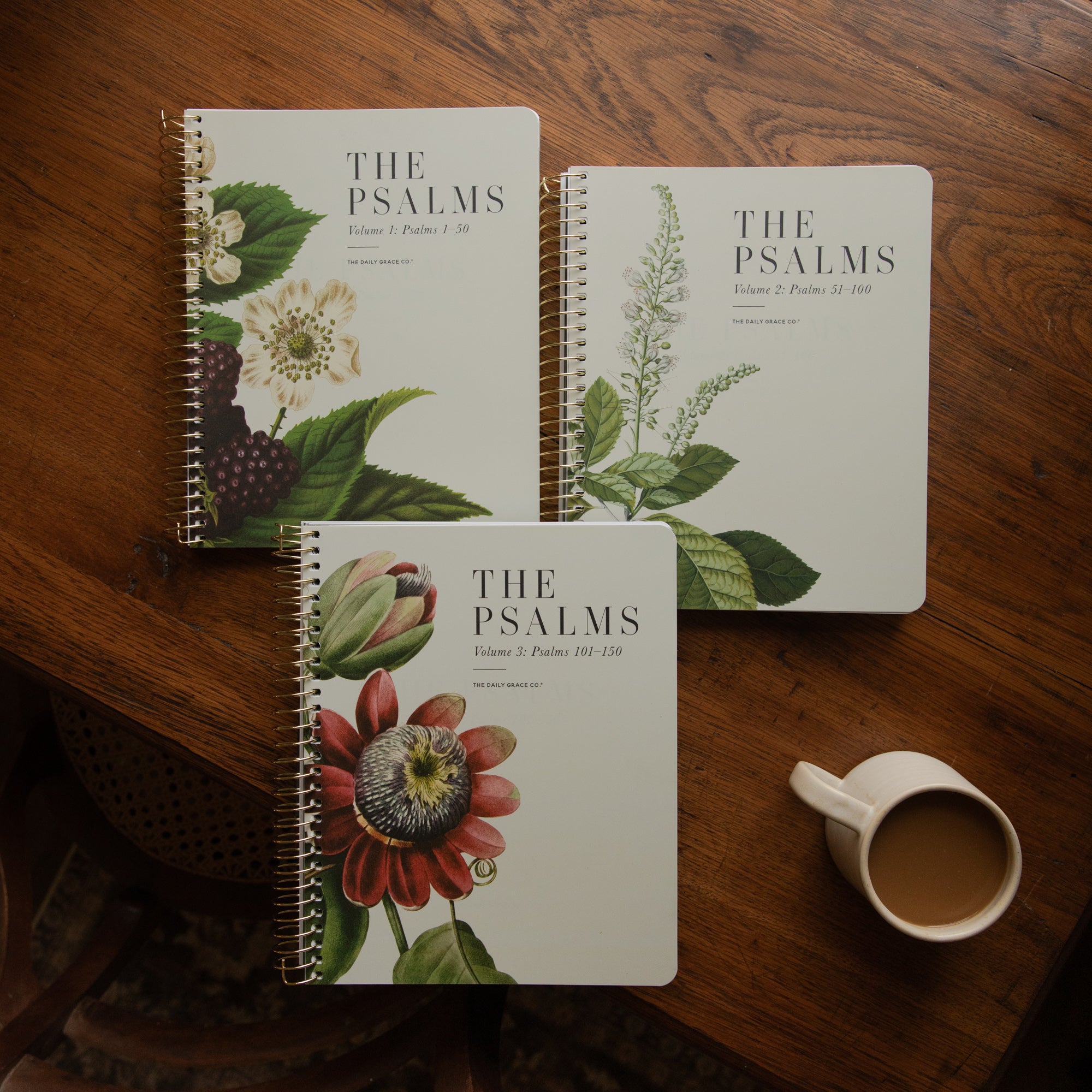 Three spiral-bound books titled "THE PSALMS" arranged on a wooden table, each with different botanical illustrations on the cover, plus a white mug of coffee nearby.