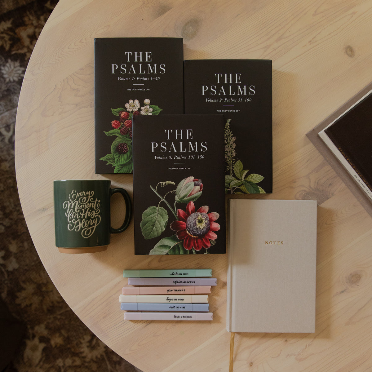 Three hardcover books titled 'The Psalms' arranged on a round light wood table, alongside a dark green ceramic mug with gold lettering, a stack of small pastel notebooks, and a cream-colored journal.