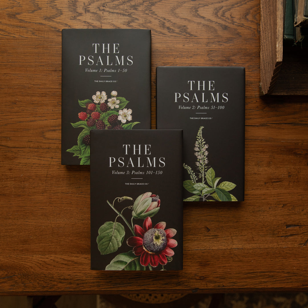 Three hardcover books titled "The Psalms" arranged on a wooden tabletop. The covers are dark charcoal/black with white serif text and botanical illustrations in red, pink, green, and white.
