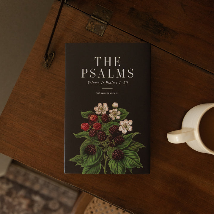 A book titled "The Psalms Volume 1: Psalms 1-50" by The Daily Grace Co. is displayed on a dark wooden table next to a white ceramic mug.