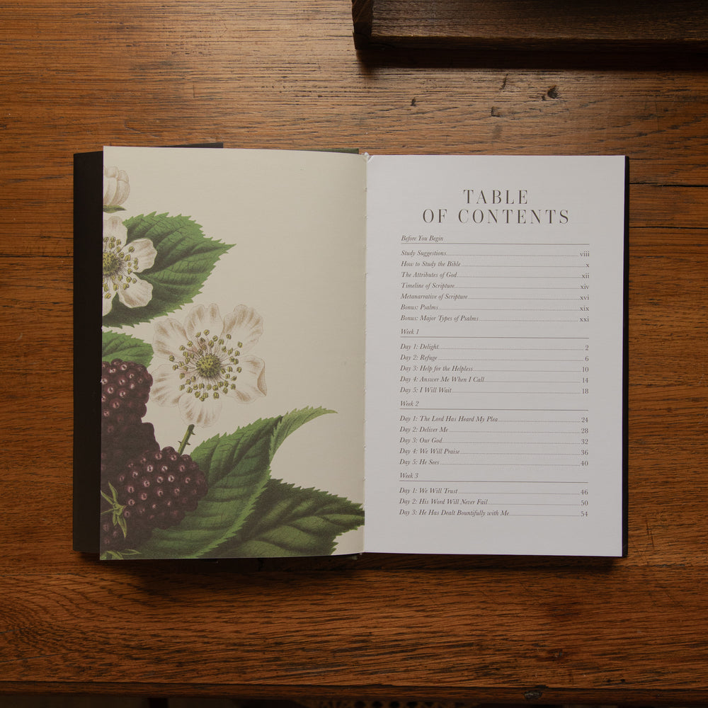 An open book laid flat on a wooden table, showing a table of contents page with a botanical illustration of white flowers and dark berries on the left page.