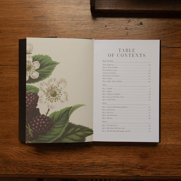 An open book laid flat on a wooden table, showing a table of contents page with a botanical illustration of white flowers and dark berries on the left page.