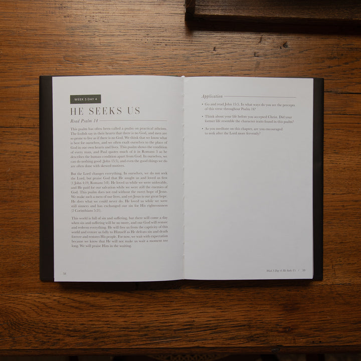 An open Bible book laid flat on a dark wooden table, showing two interior pages with printed text. The left page has the heading 'BIBLE' and the main heading 'HE SEES US' with body text below.