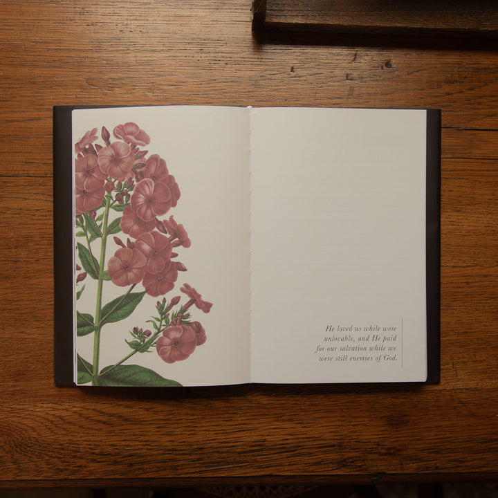 An open book or journal lying on a wooden table, showing a floral illustration with pink flowers and green leaves on the left page and a small block of text on the right page.