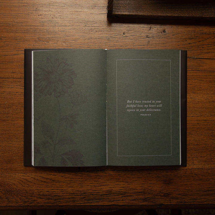 An open book with dark green pages lies on a wooden table. The left page has a subtle floral pattern, and the right page contains a small quote bordered by a thin line.