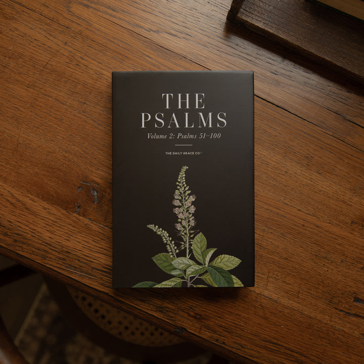 The Psalms Volume 2: Psalms 31-100 book cover by The Daily Book Club, featuring a dark charcoal cover with white text and a botanical illustration of green leaves and a flower, resting on a rustic wooden table.