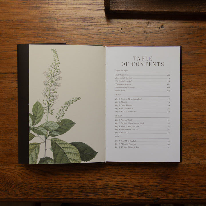 An open book showing printed table-of-contents pages on a dark wood tabletop. with botanical images on the left side of the page on a wooden table