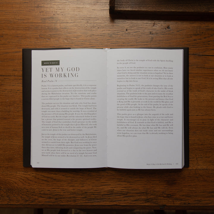 An open book lying flat on a dark wooden table, showing two printed pages with the headline 'YET MY GOD IS WORKING' and body text. No person or animal is visible.
