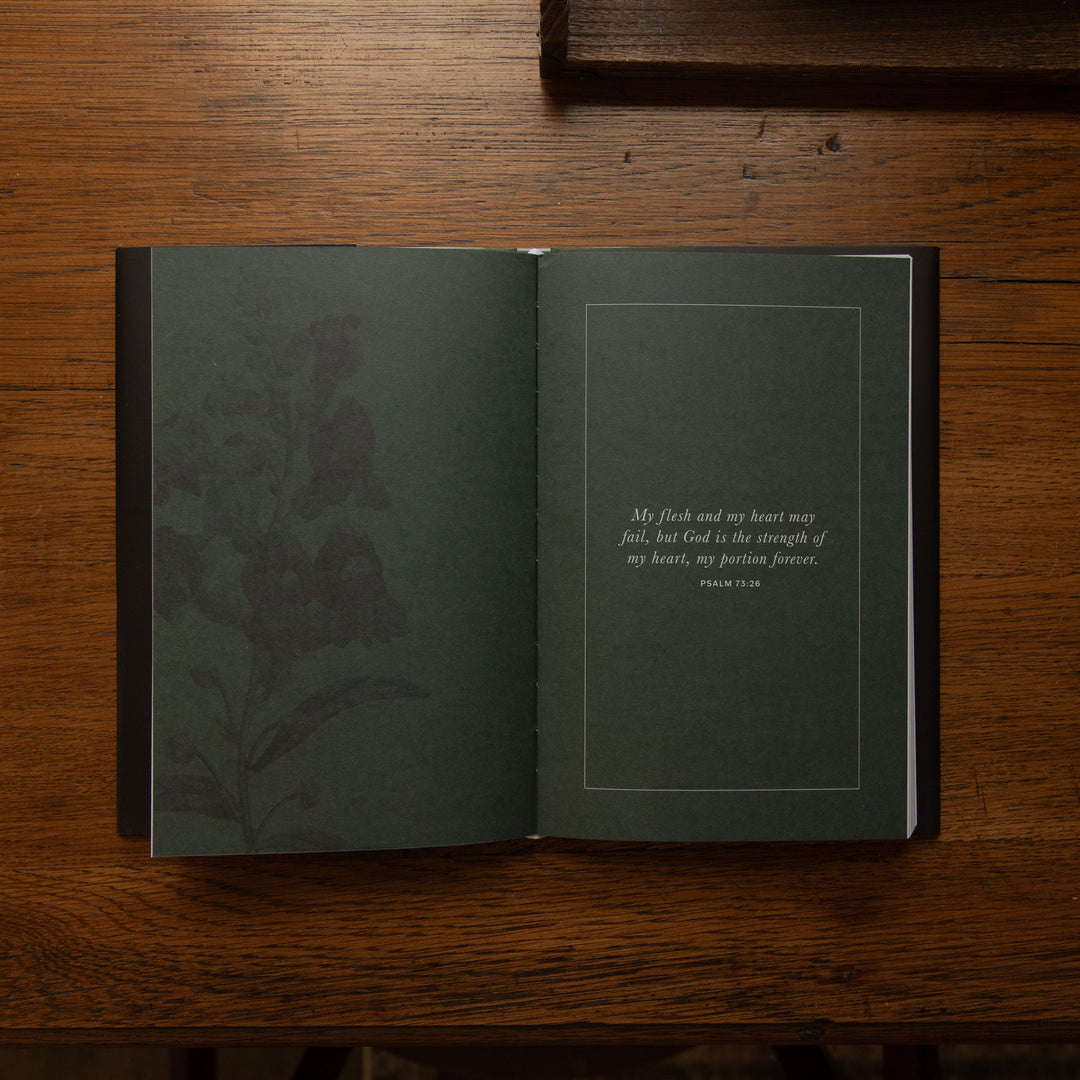 An open hardcover book with dark green pages lies on a wooden table. The left page features a subtle botanical illustration, and the right page contains a quote in white text inside a thin rectangular border.
