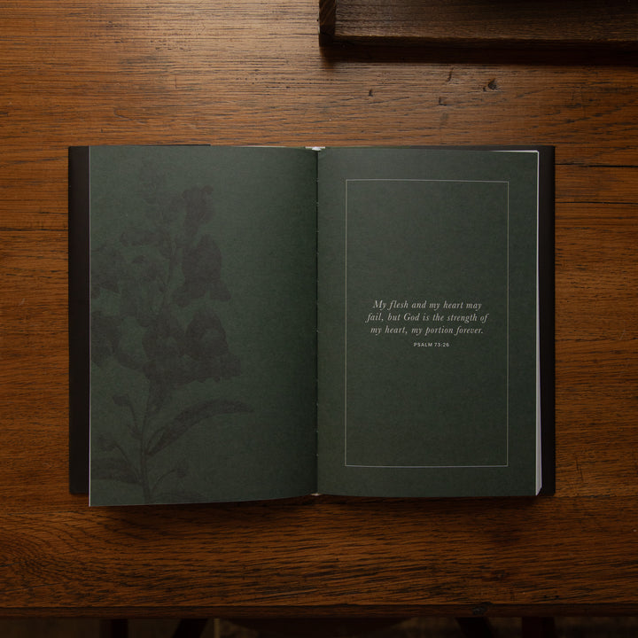 An open hardcover book with dark green pages lies on a wooden table. The left page features a subtle botanical illustration, and the right page contains a quote in white text inside a thin rectangular border.