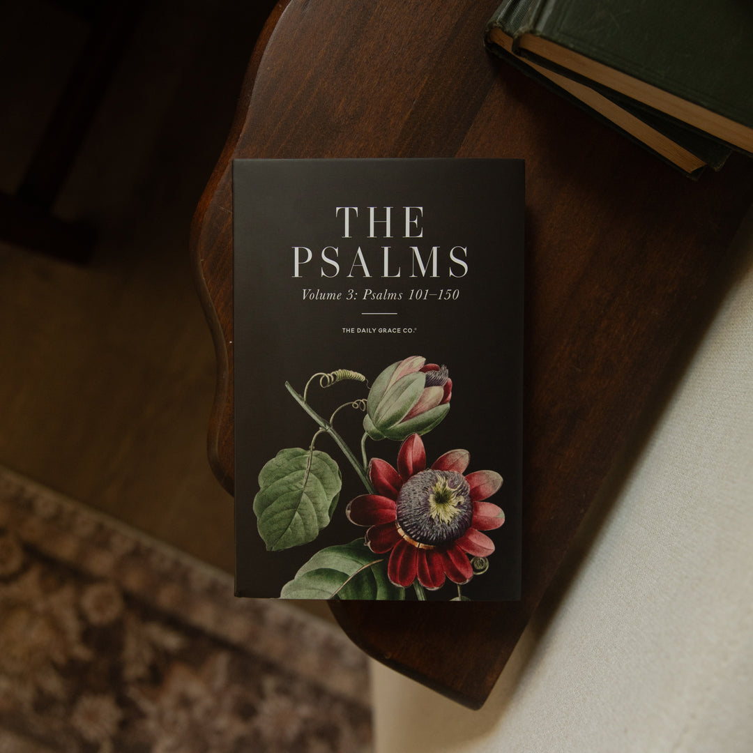 A book titled 'THE PSALMS Volume 3: Psalms 101-150' by The Daily Grace Co., featuring a black cover with white text and a botanical illustration of a red and green flower, resting on a dark wooden table.