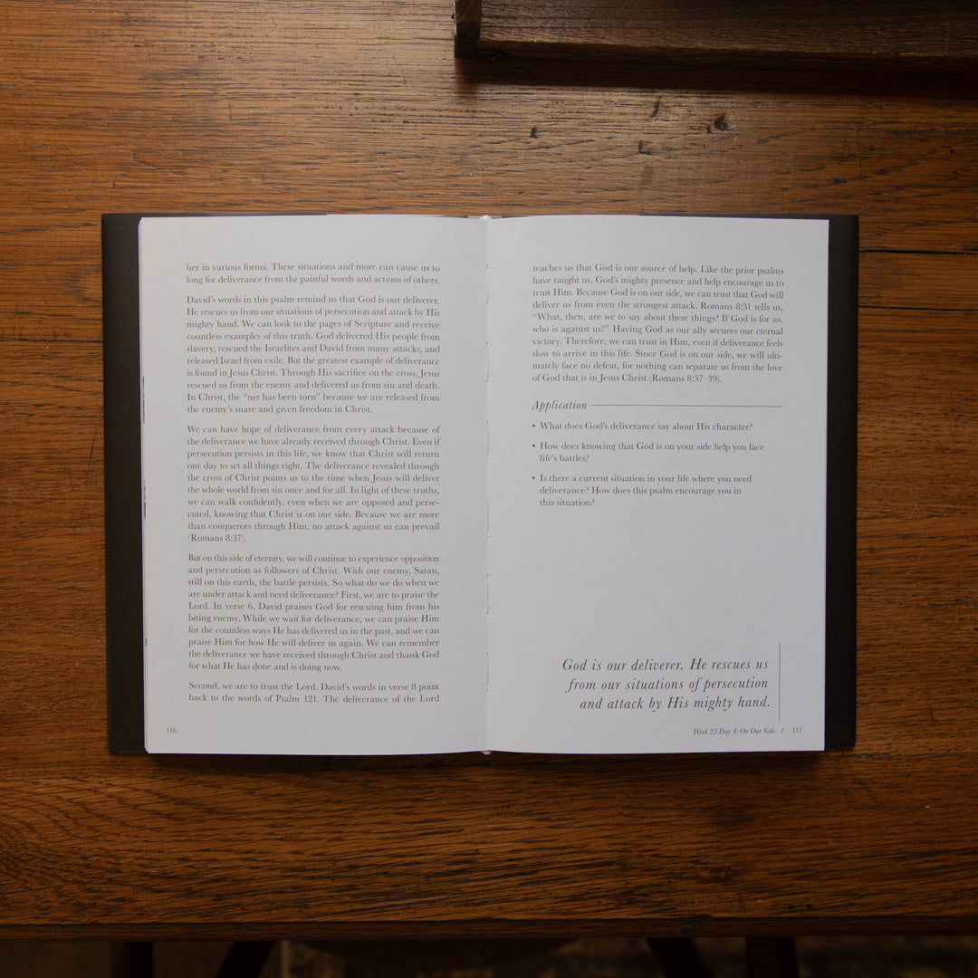 An open Bible book laid flat on a dark wooden table, showing two open pages with printed text and a small prayer at the bottom right of the right page.