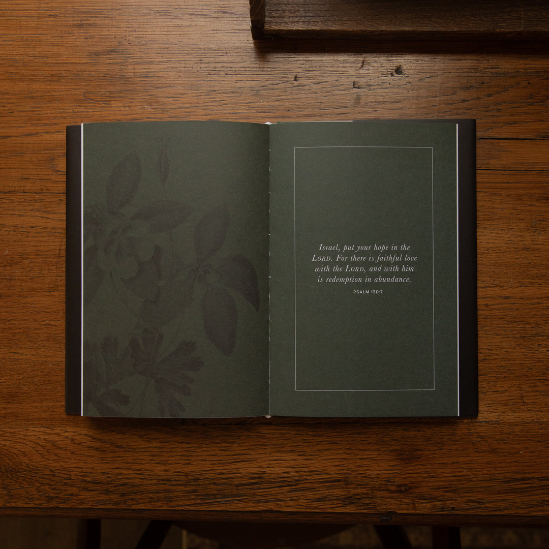 An open book lying on a wooden table, showing a dark green interior spread with a subtle floral pattern on the left page and a Bible verse from Philippians 4:13 printed on the right page.