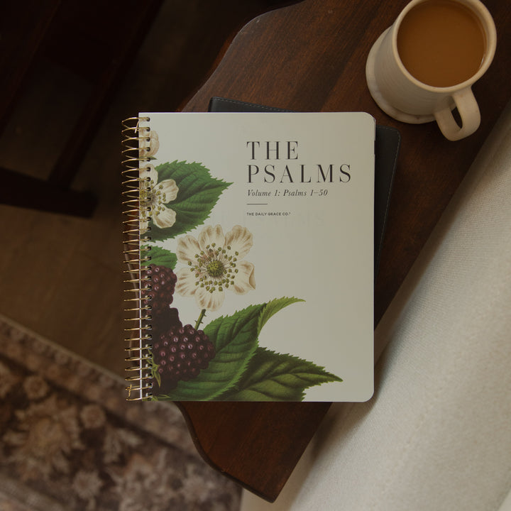 A spiral-bound notebook titled "THE PSALMS" with botanical cover art of white flowers and dark berries, placed on a dark wooden table next to a cup of coffee.
