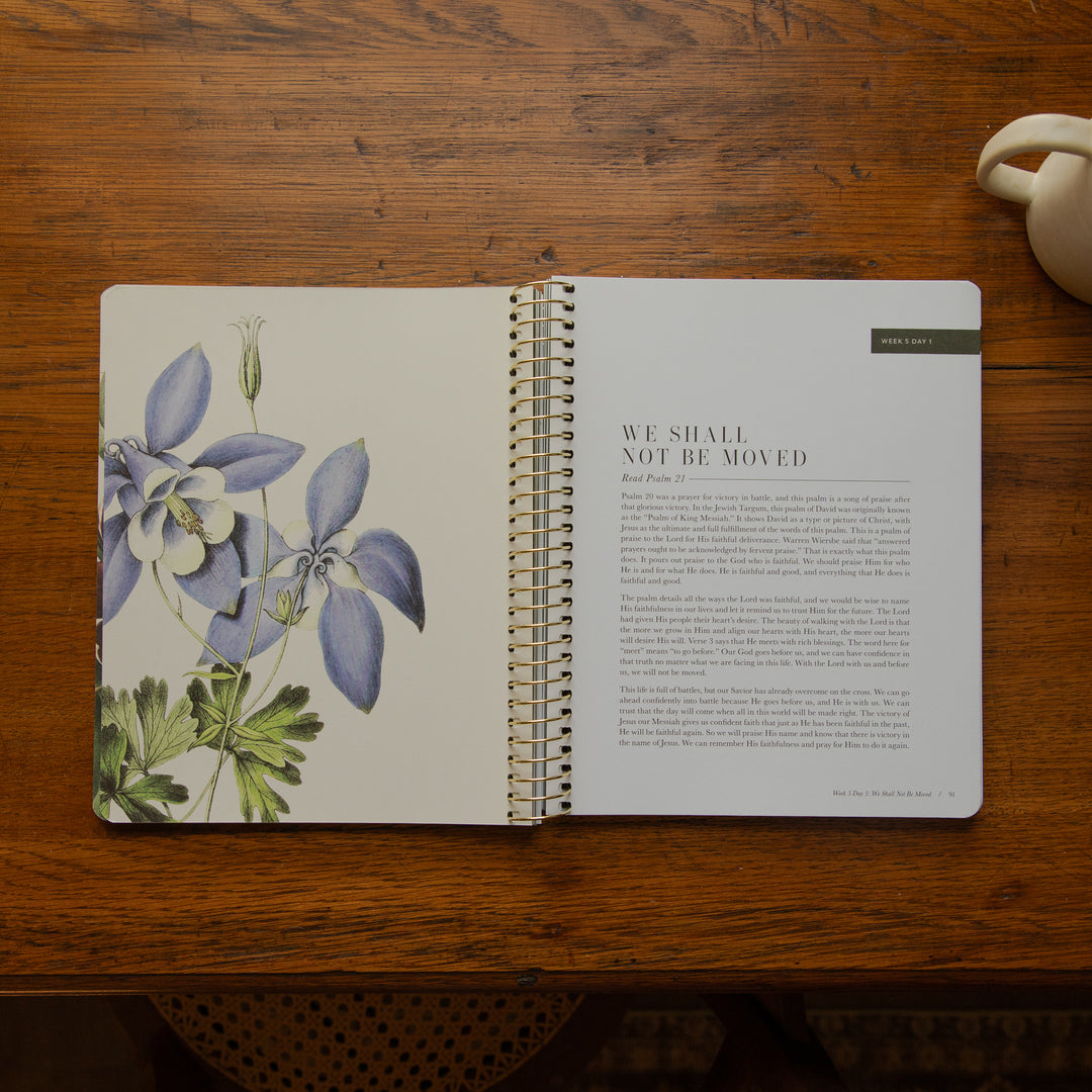 An open spiral-bound journal or notebook laid on a wooden table, showing a botanical illustration of blue and white flowers on the left page and a quote on the right page. A small label at the top right of the right page reads 'JUNE 2025'.