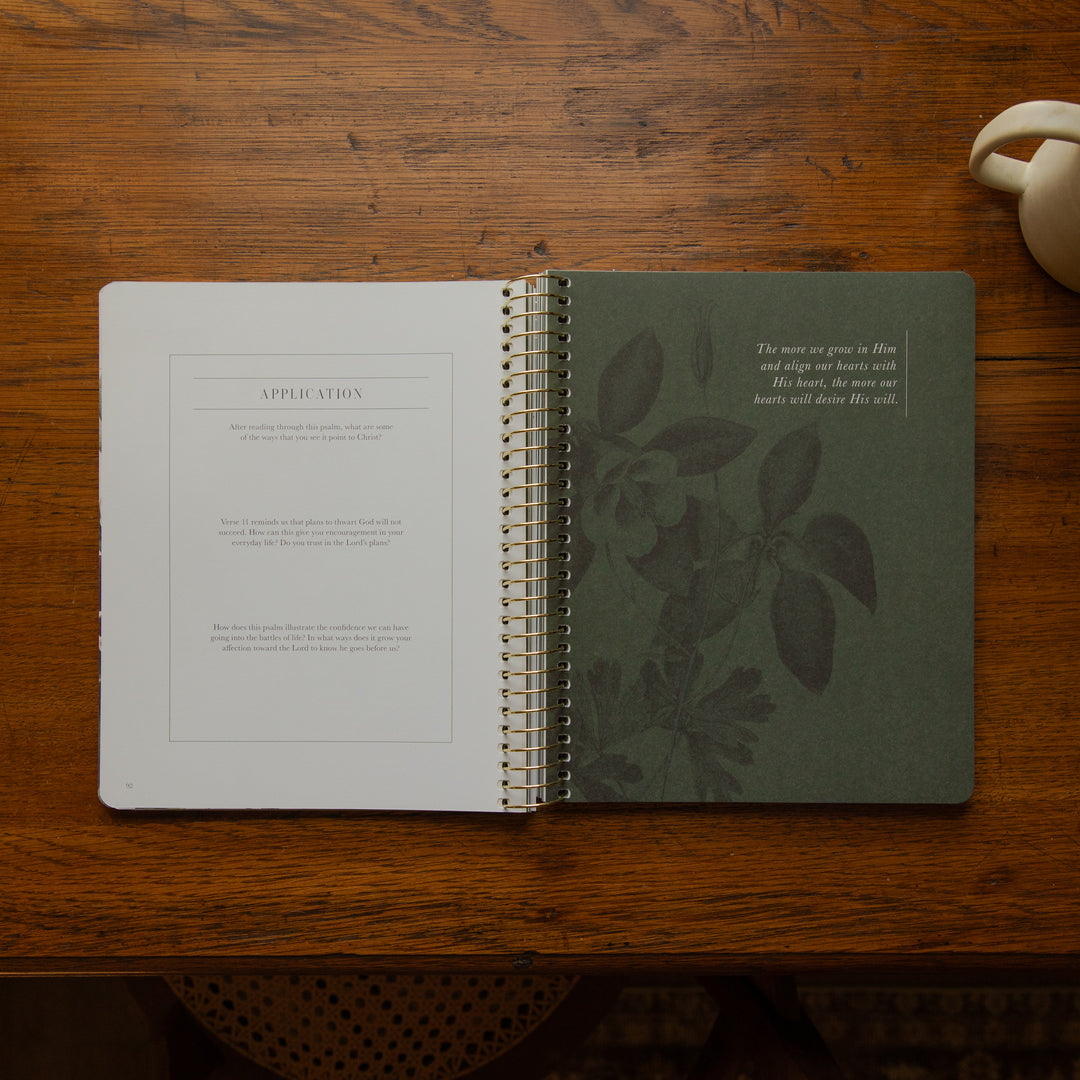 An open spiral-bound journal or notebook laid on a wooden table, showing a white left page with printed text and a dark green right page with botanical leaf illustrations and a small quote. A ceramic cup is partially visible at the top right.