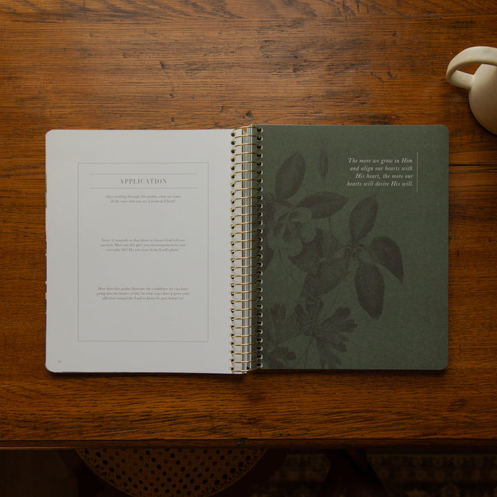 An open spiral-bound journal or notebook laid on a wooden table, showing a white left page with printed text and a dark green right page with botanical leaf illustrations and a small quote. A ceramic cup is partially visible at the top right.