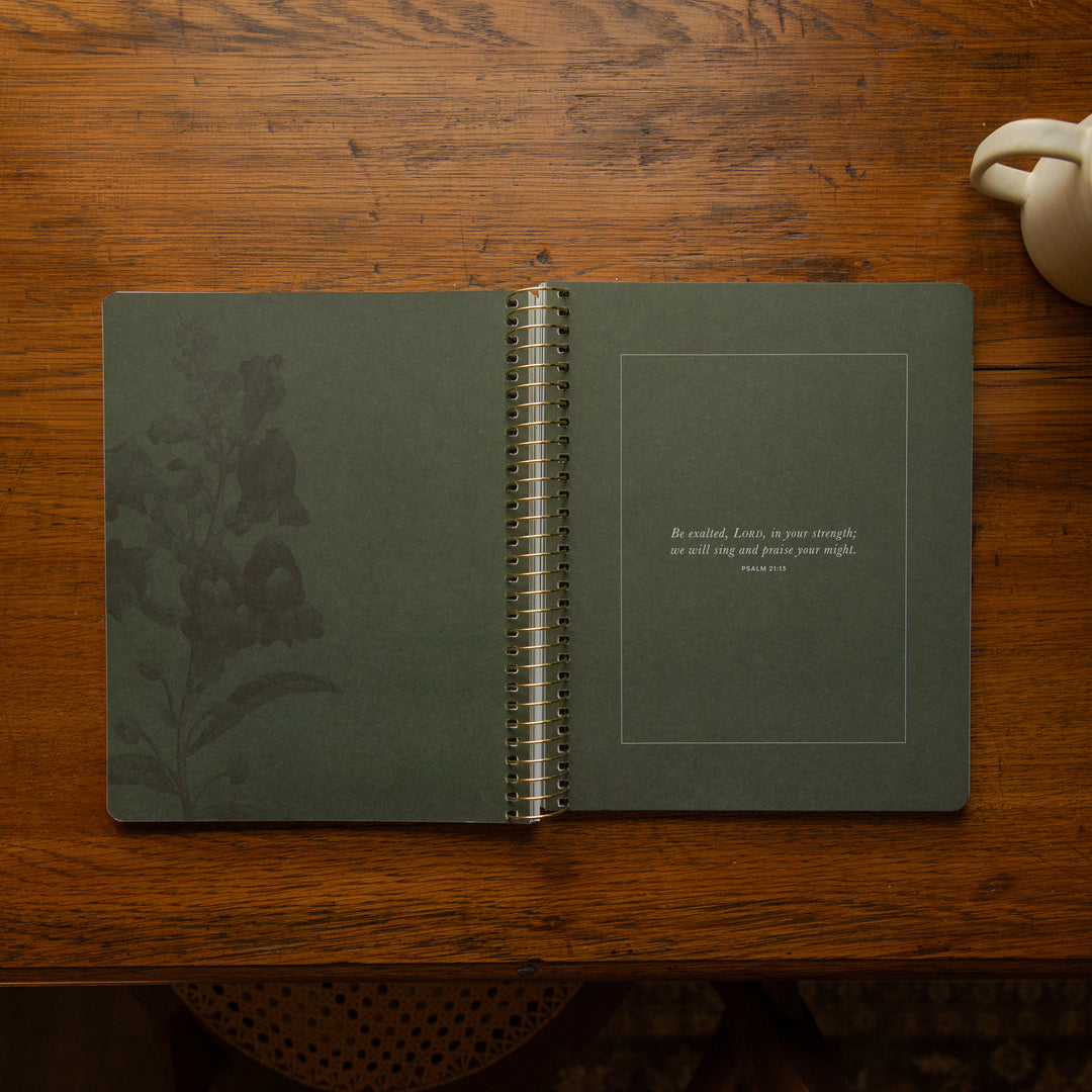 An open spiral-bound journal lies on a wooden table. The cover is a muted dark green with a subtle embossed floral design on the left page and a small quote printed on the right page inside a thin border.