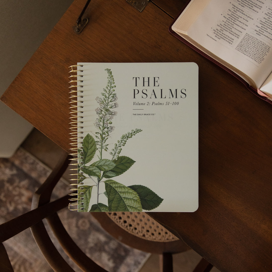 A spiral-bound notebook titled "THE PSALMS Volume 2 Psalms 31-100" with a botanical green leaf illustration on the cover, placed on a wooden table next to an open book.