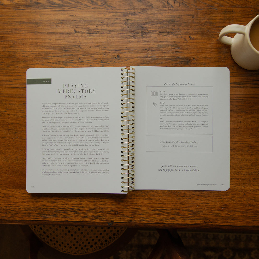 An open spiral-bound book titled 'Praying Imperative Psalms' lies on a wooden table, with a cream ceramic mug partially visible in the top right corner.