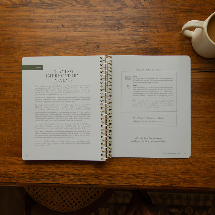 An open spiral-bound book titled 'Praying Imperative Psalms' lies on a wooden table, with a cream ceramic mug partially visible in the top right corner.
