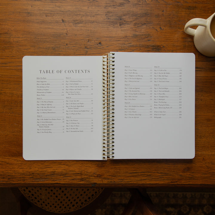An open spiral-bound notebook showing printed table of contents pages, placed on a wooden table with a cream mug partially visible in the top right corner.