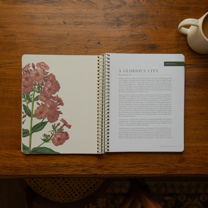 An open spiral-bound book or journal laid on a wooden table, showing a floral illustration on the left page and a text page titled 'A GLORIOUS CITY' on the right. A cream mug is partially visible in the top-right corner.