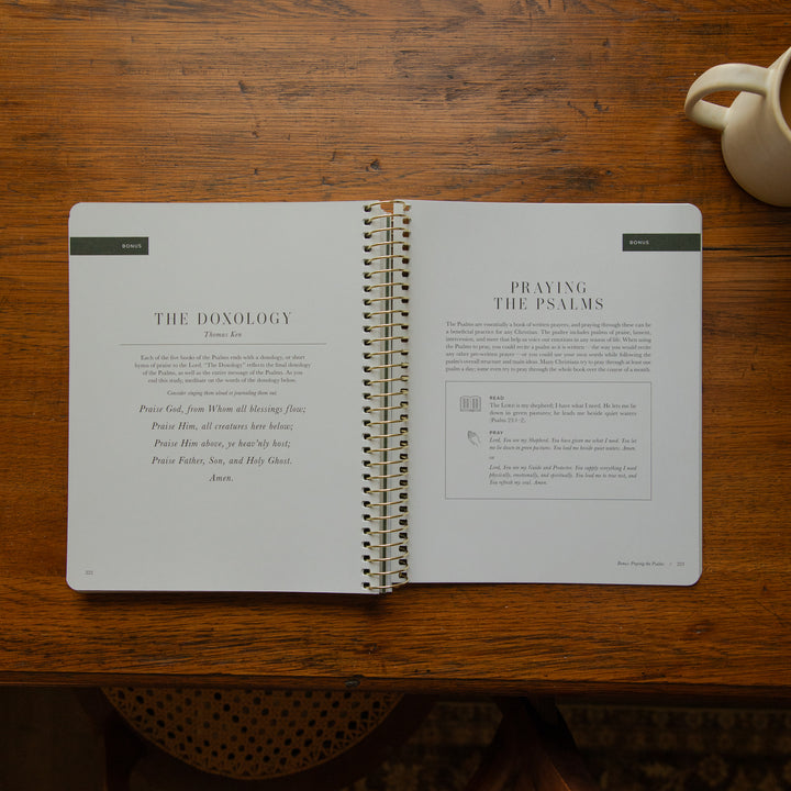 An open spiral-bound book laid flat on a wooden table, showing printed pages titled 'The Donology' and 'Praying the Psalms.' A small cream-colored ceramic cup is partially visible in the top-right corner.