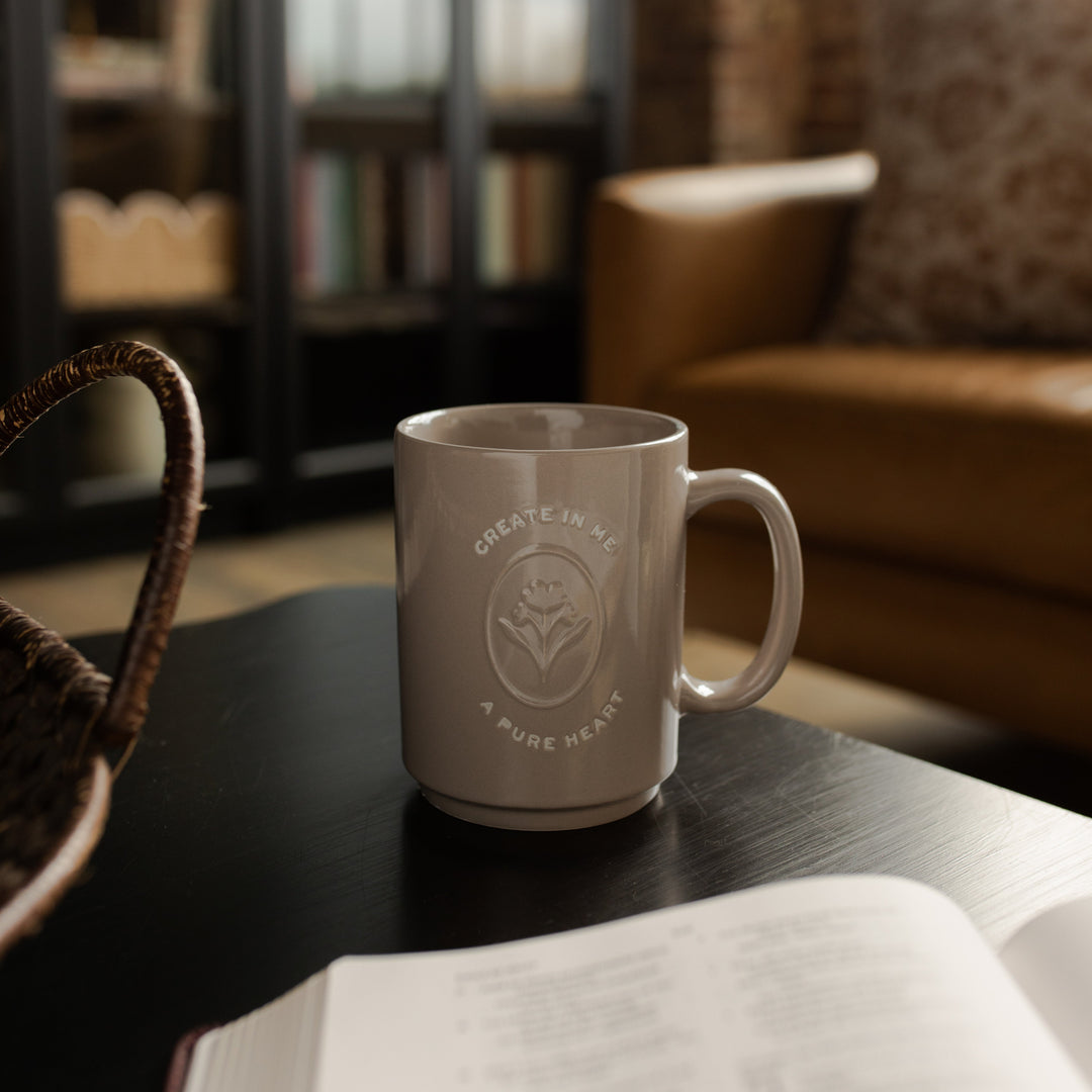 A mug with the words Create In Me A Pure Heart on a brown table next to an open bible
