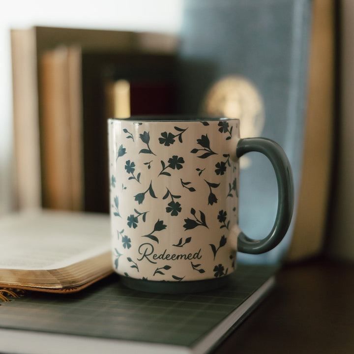 A ceramic mug with a green floral pattern on a cream background and the word 'Redeemed' printed on the front, styled on a desk with books in a warm, softly lit setting.