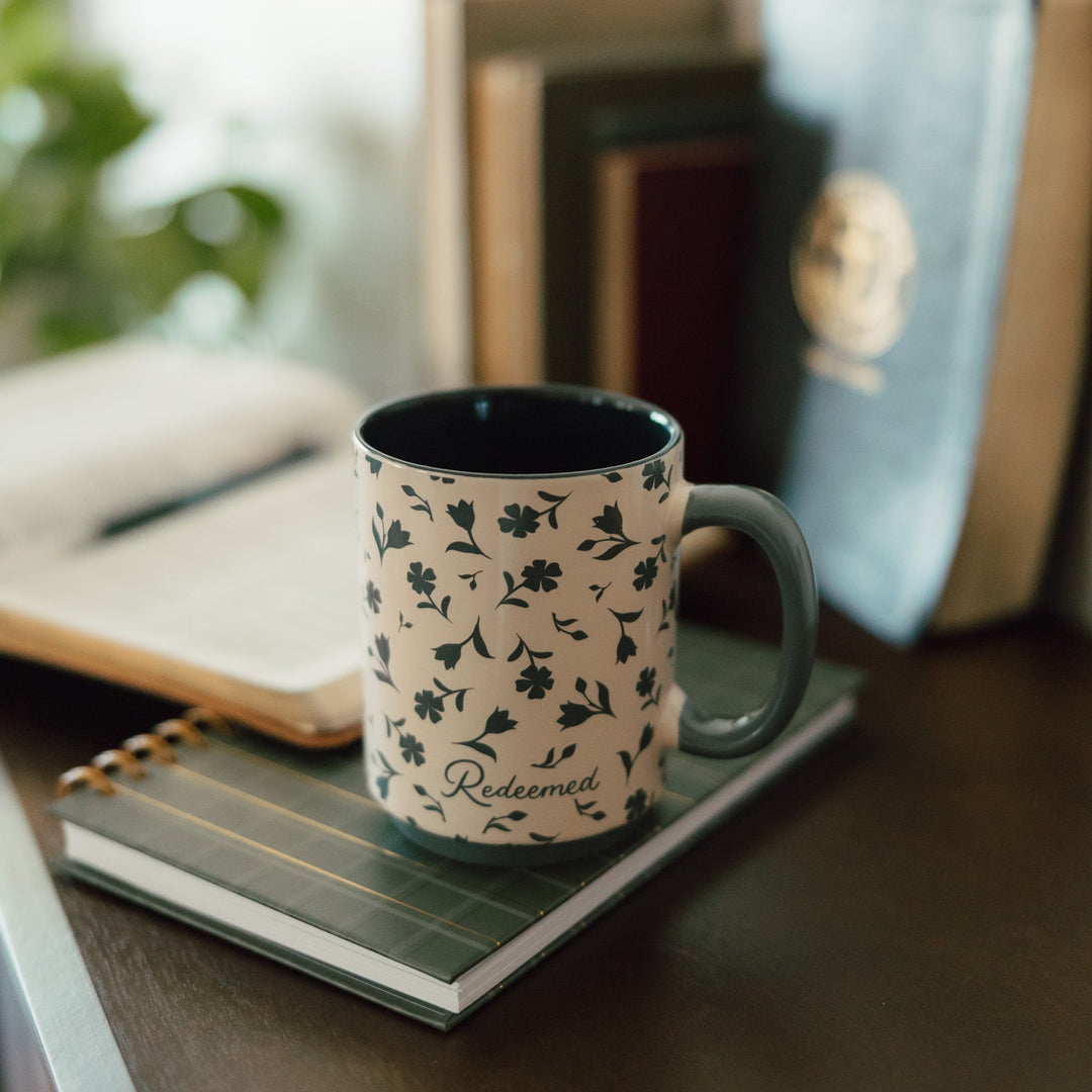 A ceramic mug with a green floral pattern on a cream background sits on a notebook on a wooden table, with books blurred in the background.