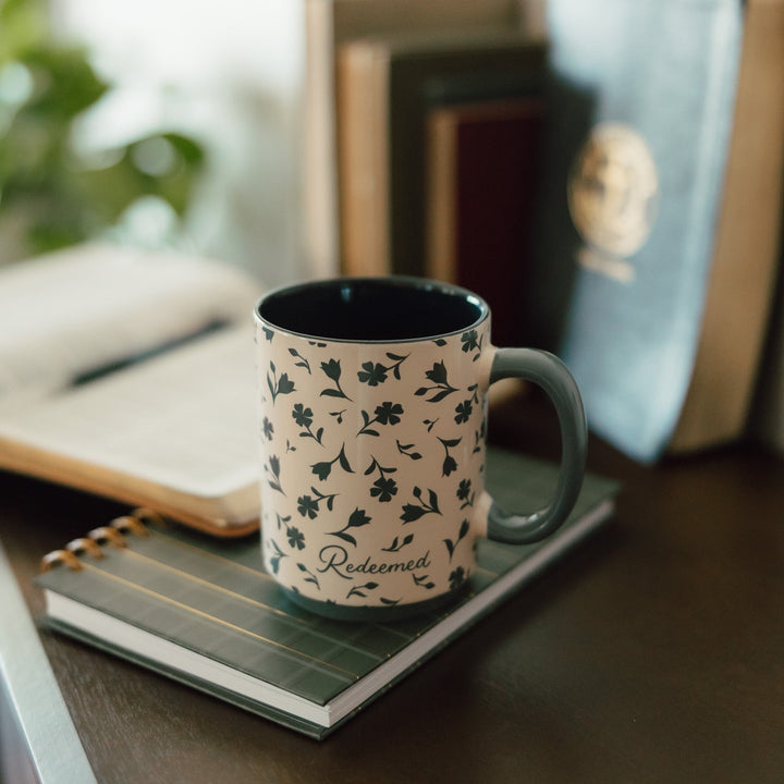 A ceramic mug with a green floral pattern on a cream background sits on a notebook on a wooden table, with books blurred in the background.