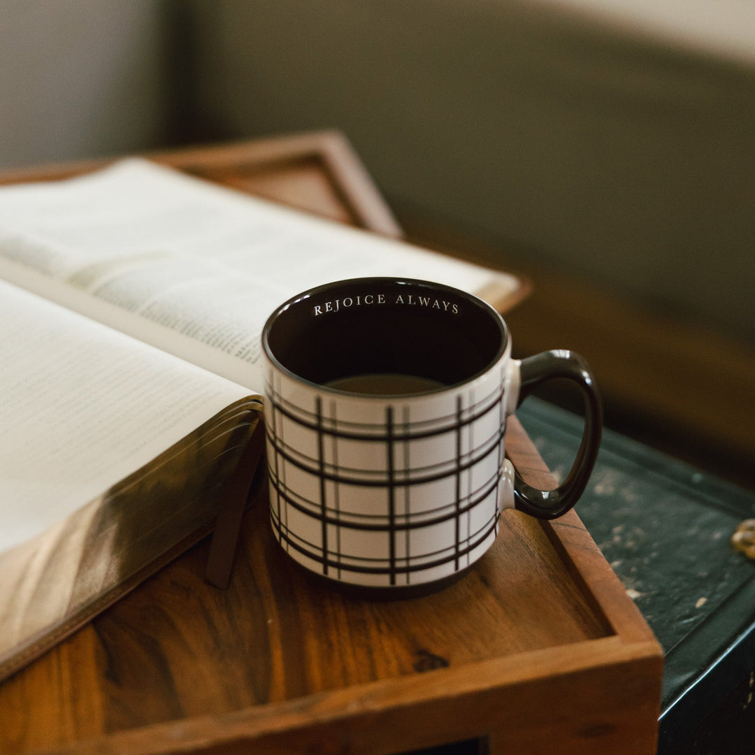A black and white plaid ceramic mug with the words 'REJOICE ALWAYS' printed inside, resting on a wooden tray next to an open book in a cozy indoor setting.