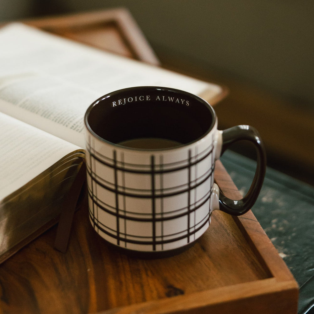 A brown and white plaid ceramic mug with the words 'REJOICE ALWAYS' printed inside, resting on a wooden tray next to an open book in a cozy indoor setting.