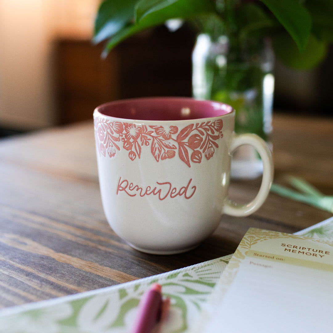 Pink Renewed Mug sitting on a table next to a notepad and green plant. 