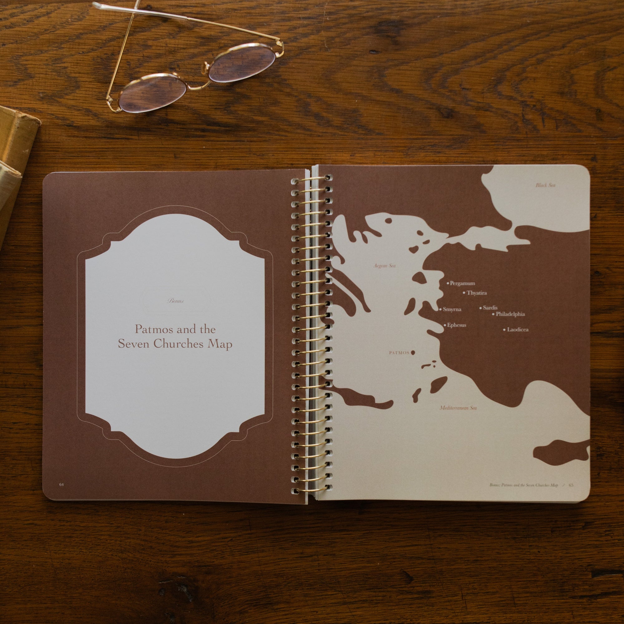 Revelation Spiral Study opened to page 64, Patmos and the seven churches map, on a wooden table next to a pair of glasses