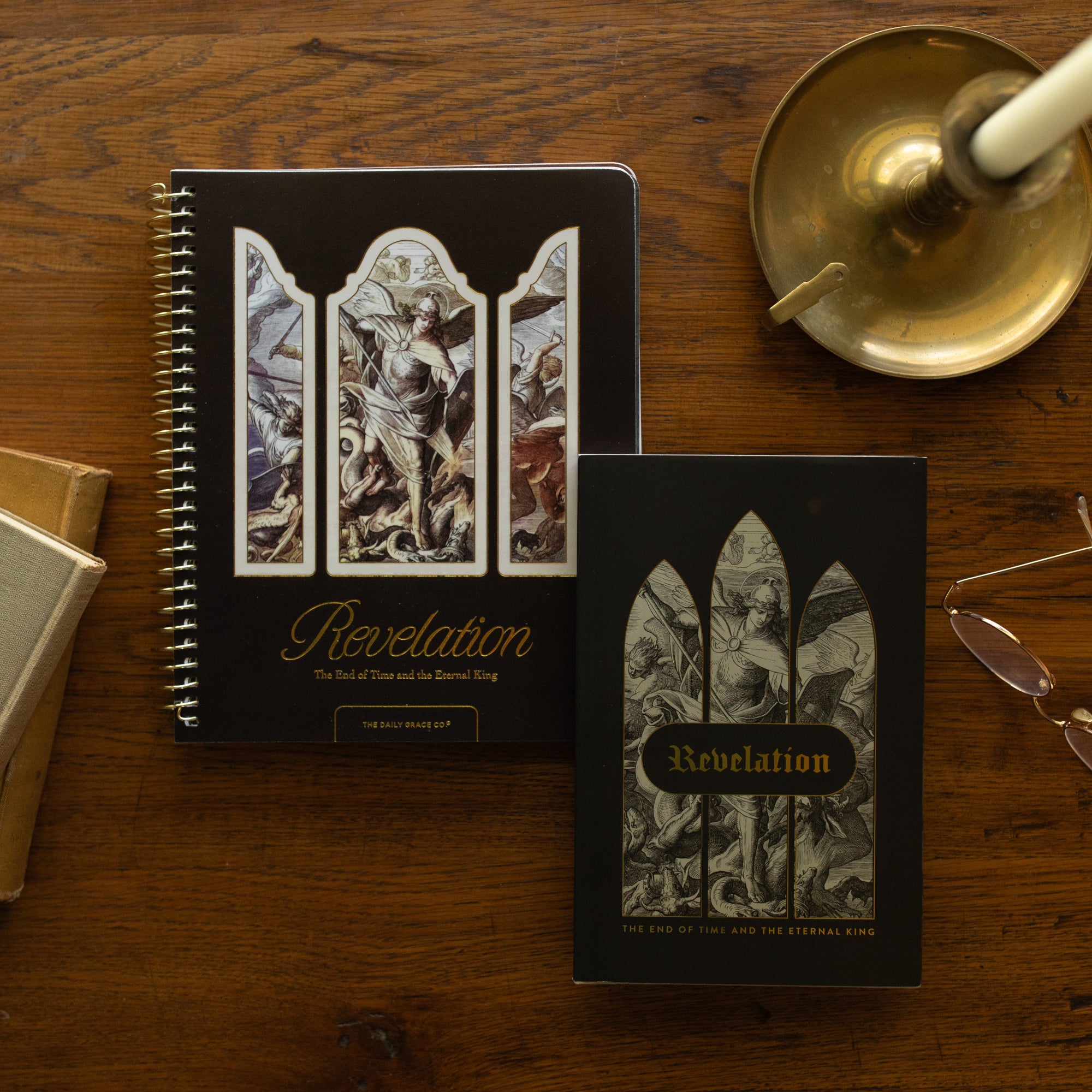Revelation Hardcover and Spiral on a wooden table next to a bronze candlestick