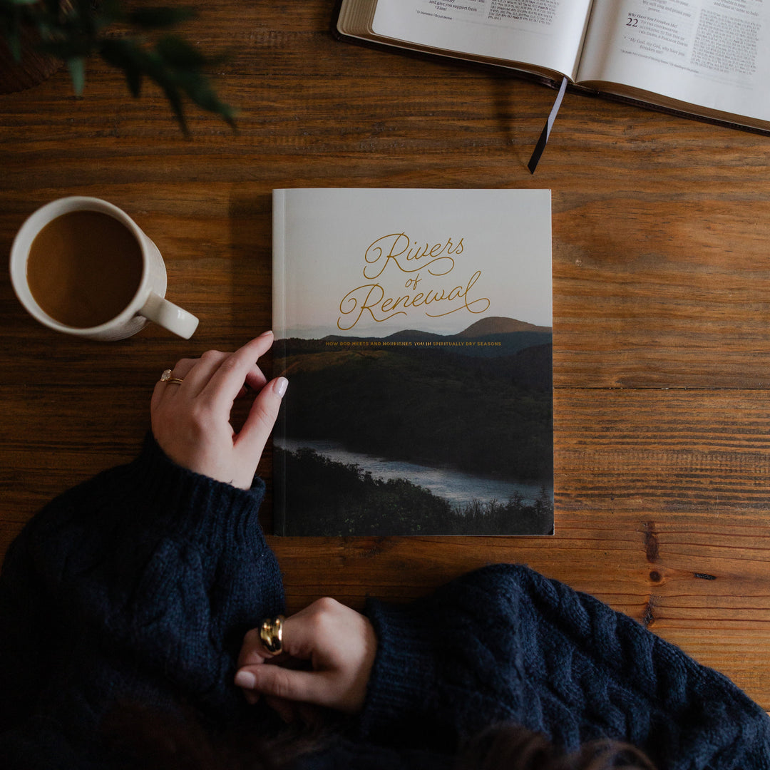 Rivers of Renewal Study cover on a brown wooden table with a woman resting on the spine of the book with a coffee next to it on a brown wooden table with an open bible on the corner