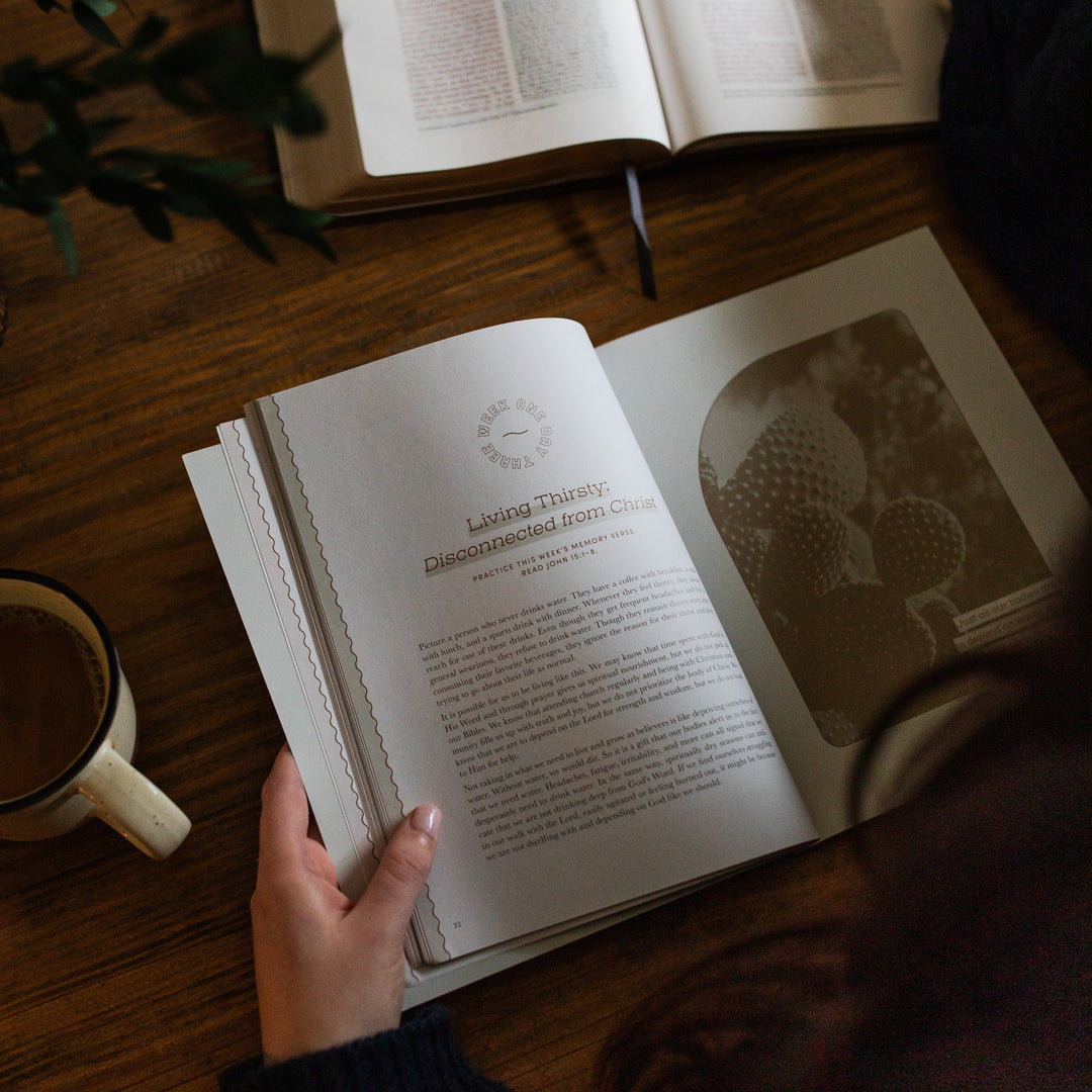 Rivers of Renewal Study opened to week one day three page laying on a brown wooden table; A woman is holding the study open; There is a coffee cup and an open bible laying on the table next to the study